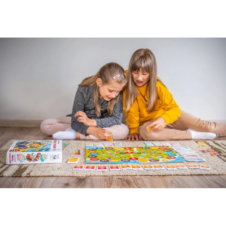 Dwie dziewczynki siedzą na dywanie i grają w grę planszową "Mały inwestor", rozkładając karty i banknoty wokół kolorowej planszy.