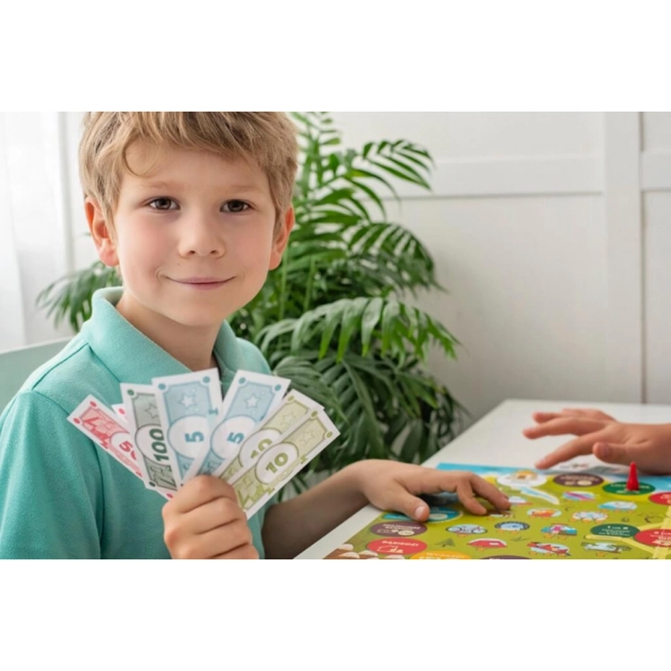 Uśmiechnięty chłopiec trzyma w dłoni wachlarz kolorowych banknotów z gry, siedząc przy stole z rozłożoną planszą.