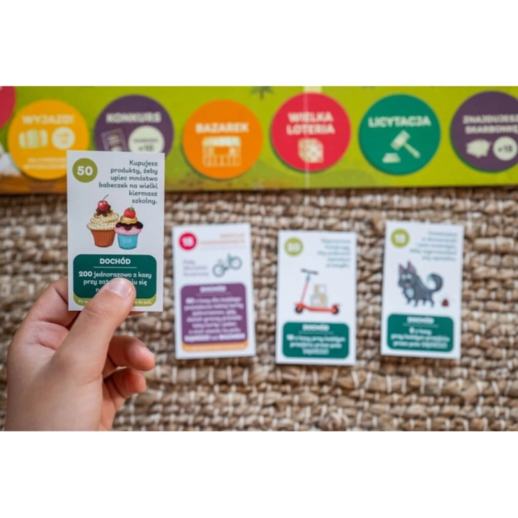 Dłoń trzymająca kartę akcji z ilustracją babeczek i opisem dochodu; w tle widoczne inne karty oraz fragment planszy.