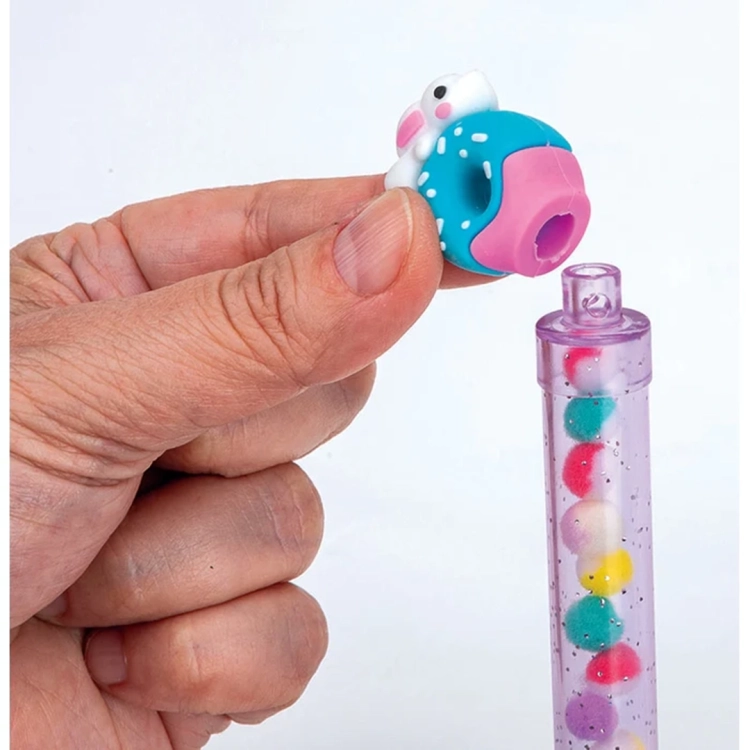 Detal dłoni nakładającej kolorową nasadkę w kształcie pączka z króliczkiem na przezroczysty korpus długopisu wypełniony pomponami.