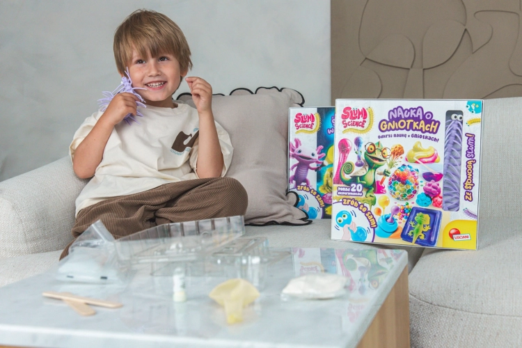 Chłopiec na kanapie trzyma fioletową stonogę; na stole przed nim leżą akcesoria do eksperymentów i pudełko zestawu.