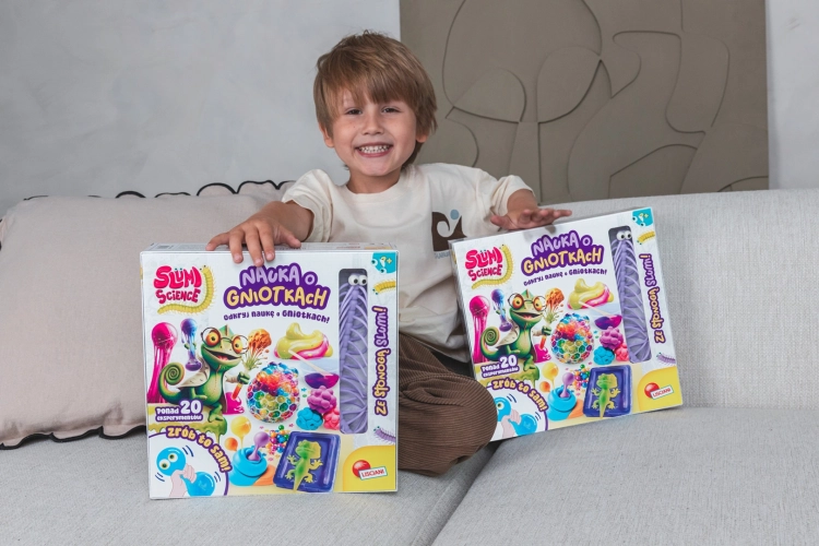 Uśmiechnięty chłopiec siedzący na jasnej kanapie, trzymający dwa duże pudełka z zestawami kreatywnymi Slumi Science.