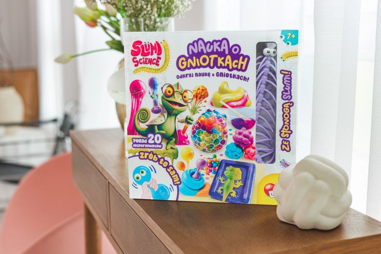 Pudełko zestawu „Nauka o gniotkach” wyeksponowane na drewnianej komodzie obok dekoracyjnej białej rzeźby i kwiatów.