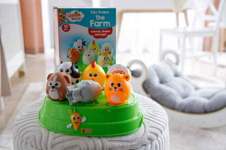 Zabawka edukacyjna Farma na miękkim pufie; na pierwszym planie zielona podstawa i kolorowe figurki, w tle pudełko zabawki.