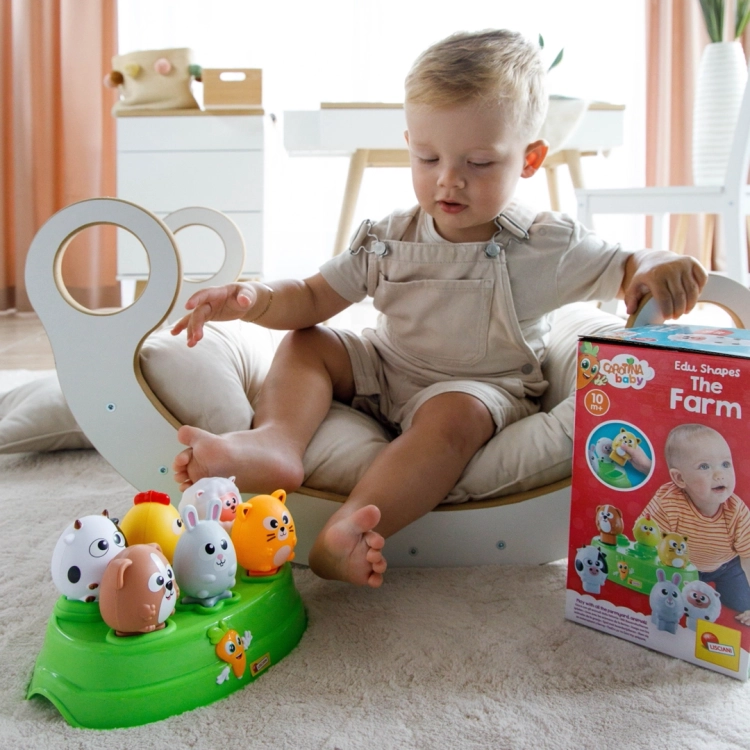 Dziecko siedzące w bujaku na dywanie bawi się sorterem edukacyjnym Farma; obok stoi kolorowe pudełko zabawki Lisciani.