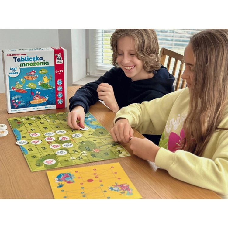 Dwoje dzieci gra przy stole w grę edukacyjną Kapitan Nauka, rozkładając żetony z żabami i krabami na kolorowej planszy z liczbami.