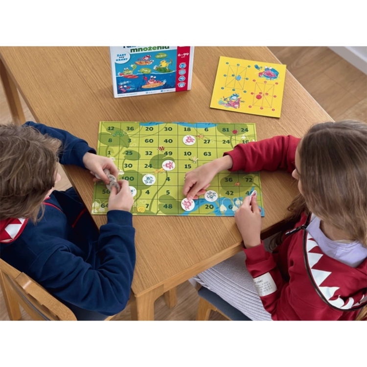 Widok z góry na dwoje dzieci grających w „Żaby czy kraby”; na stole widoczna plansza do mnożenia, żetony oraz opakowanie gry.