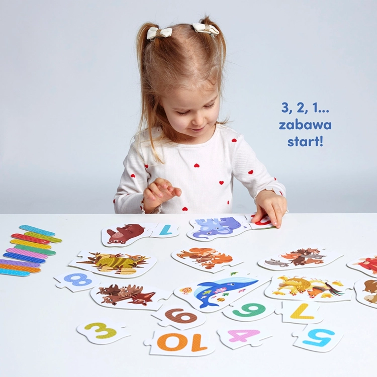 Dziewczynka siedząca przy białym stole układa dwuelementowe puzzle edukacyjne z kolorowymi zwierzętami i cyframi.