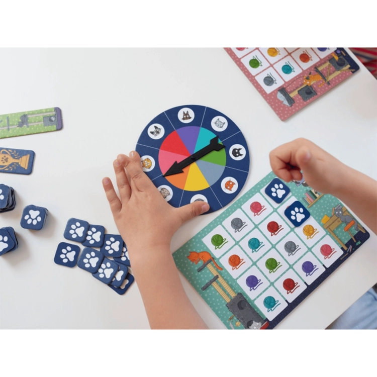 Widok z góry na ręce dziecka grającego w Bingo; na stole leży tarcza ze strzałką, plansze z kolorowymi kłębkami wełny i żetony z łapkami.