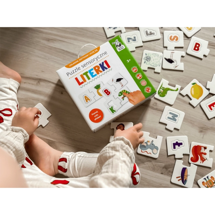 Dziecko siedzące na podłodze układa dwustronne puzzle edukacyjne z literami alfabetu i wesołymi ilustracjami zwierząt oraz przedmiotów.