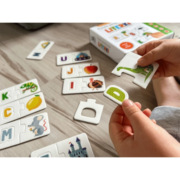 Dłonie dziecka dopasowującego element z literą „D” do obrazka z dinozaurem; w tle inne ułożone zestawy, np. jabłko, indyk, mysz.