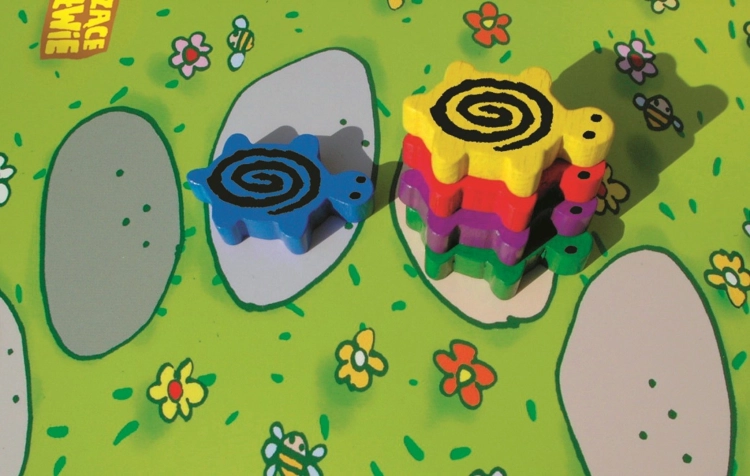 Zbliżenie na drewniane figurki żółwi na planszy: niebieski żółw stoi obok słupka złożonego z żółtego, czerwonego, fioletowego i zielonego żółwia.