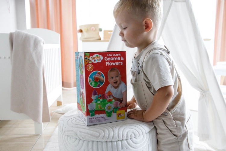 Dziecko stojące przy miękkim pufie, na którym ustawione jest duże czerwone opakowanie zabawki Carotina Baby.