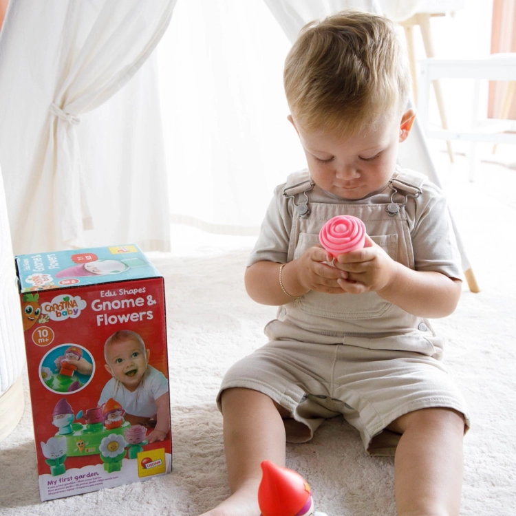 Dziecko siedzące na jasnym dywanie trzyma w rączkach różową figurkę kwiatuszka; obok stoi pudełko sortera.