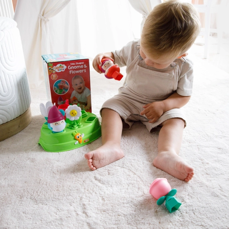Dziecko bawiące się zestawem na dywanie; podnosi figurkę krasnala, próbując dopasować ją do otworu w zielonej podstawie.