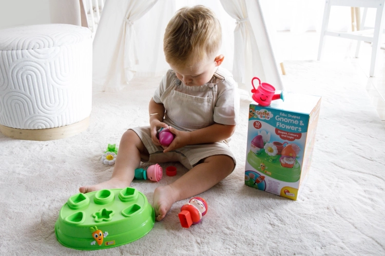 Dziecko bawiące się elementami zestawu Carotina Baby na dywanie; obok widoczna zielona podstawa sortera i opakowanie.