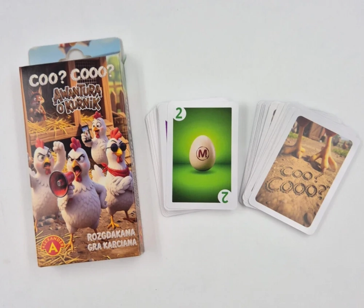 Opakowanie gry „Awantura o kurnik” obok dwóch stosów kart. Jedna karta pokazuje jajko z literą M na zielonym tle, druga tył karty z kurzymi łapkami.
