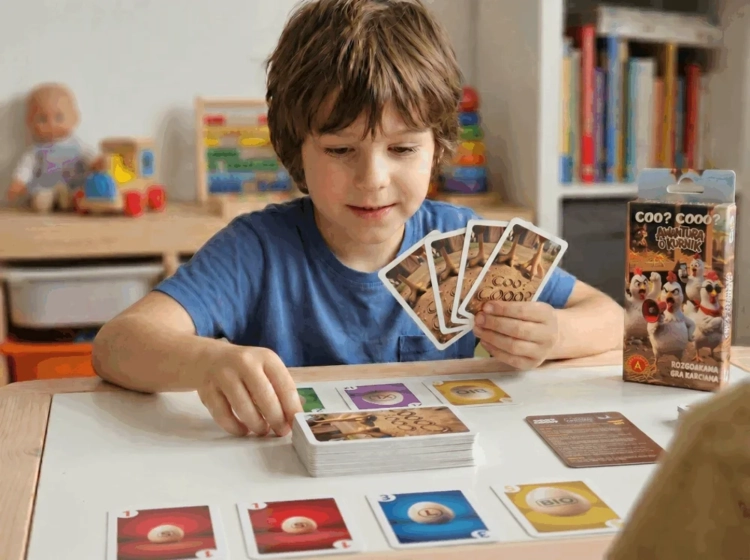 Chłopiec siedzi przy stole i trzyma w dłoniach karty, grając w „Awanturę o kurnik”. Na blacie rozłożone są stosy kart oraz instrukcja gry.