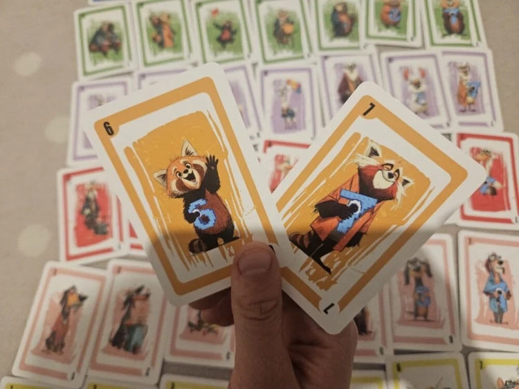 Dłoń trzymająca dwie pomarańczowe karty z numerami 6 i 7. Na kartach widnieją ilustracje pandy małej – jedna macha łapką, a druga trzyma niebieski klocek z liczbą 7.