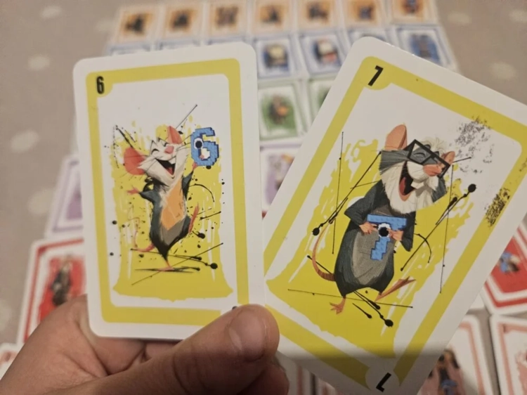 Dwie żółte karty trzymane w dłoni, przedstawiające radosne białe myszki z niebieskimi elementami liczb 6 i 7 na tle rozłożonych innych kart z gry.