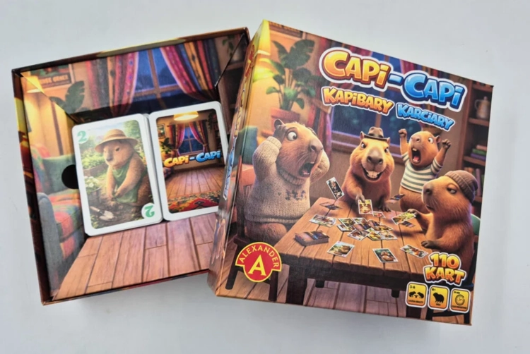 Otwarte pudełko gry prezentujące wnętrze z nadrukiem pokoju. W środku znajdują się dwie talie kart z ilustracjami kapibar.