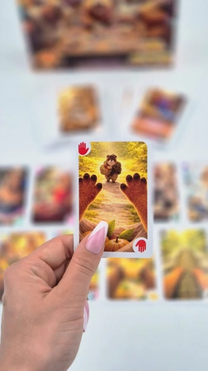 Dłoń trzymająca jedną kartę z gry na rozmytym tle pozostałych elementów. Karta przedstawia kapibarę idącą ścieżką i jest oznaczona symbolem czerwonej dłoni (STOP).