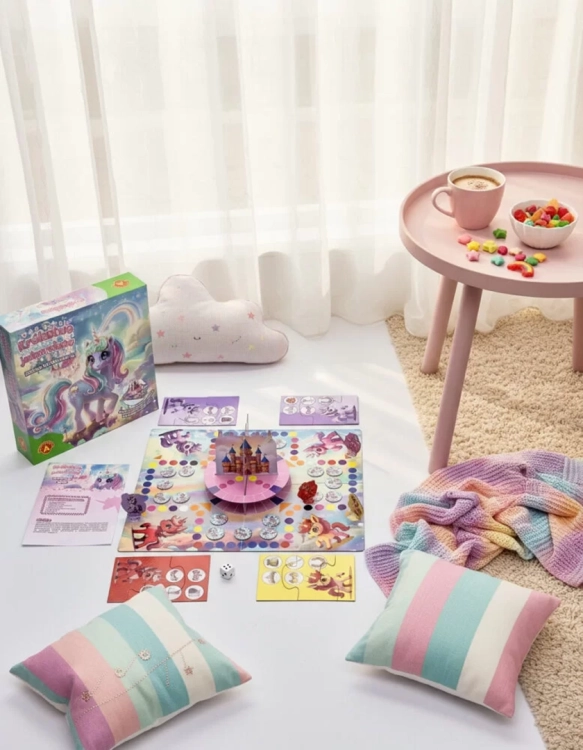 Aranżacja gry w jasnym wnętrzu. Obok planszy leżą poduszki, kolorowy koc oraz stolik z kawą i miseczką słodyczy.