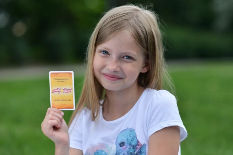 Dziewczynka siedzi na trawie i pokazuje do kamery kartę z pytaniem z gry „Gorący Ziemniak Junior”.