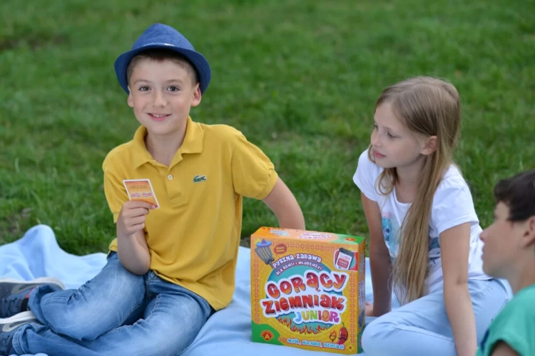 Troje dzieci siedzi na kocu w parku obok pudełka gry „Gorący Ziemniak Junior”. Chłopiec w niebieskim kapeluszu trzyma kartę z pytaniem.