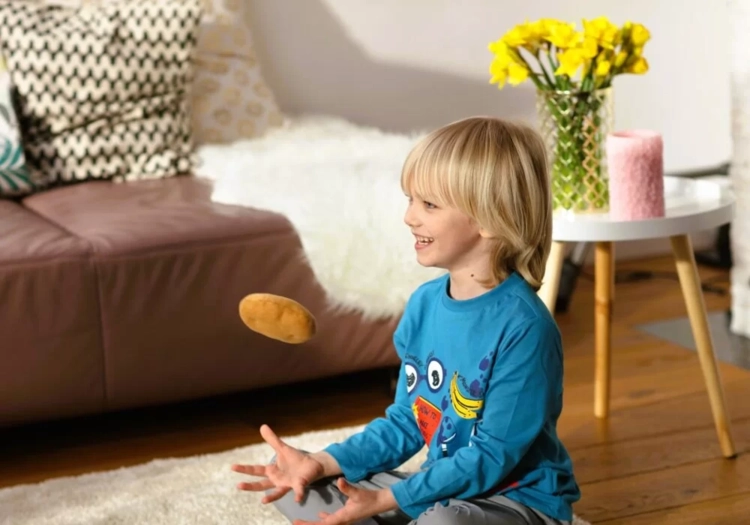 Chłopiec siedzący na dywanie rzuca w górę pluszowego ziemniaka, świetnie się bawiąc.