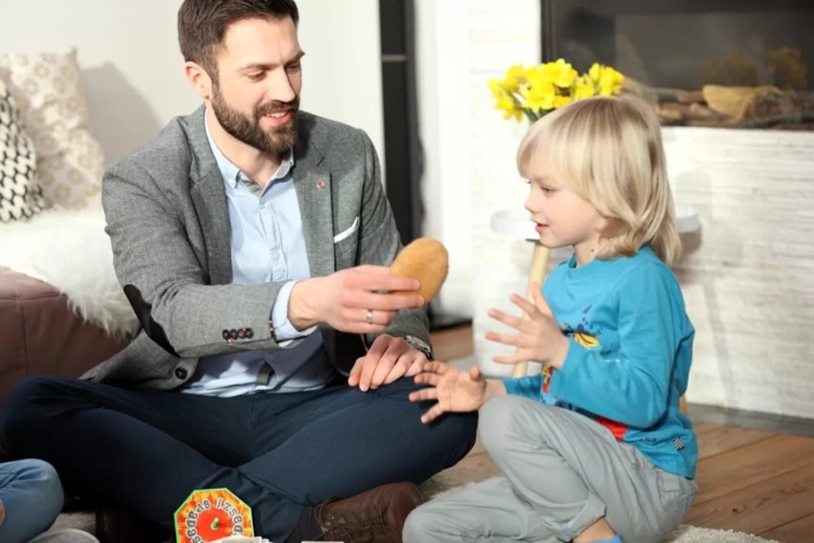 Mężczyzna i chłopiec siedzą na podłodze i grają w Gorącego Ziemniaka, podając sobie pluszaka.