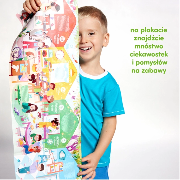 Roześmiany chłopiec chowa się za dużym, pionowym plakatem edukacyjnym, który jest częścią zestawu. Na plakacie widać kolorowe sale przedszkolne.