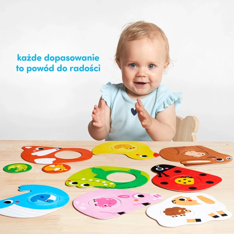 Uśmiechnięte dziecko klaszcze w dłonie przy stole zastawionym puzzlami ze zwierzątkami. Napis: każde dopasowanie to powód do radości.