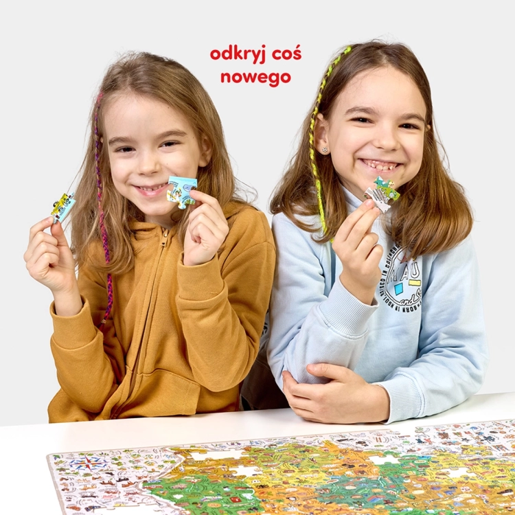 Dwie uśmiechnięte dziewczynki prezentują do kamery pojedyncze elementy puzzli na tle rozłożonej układanki Mapa Polski.