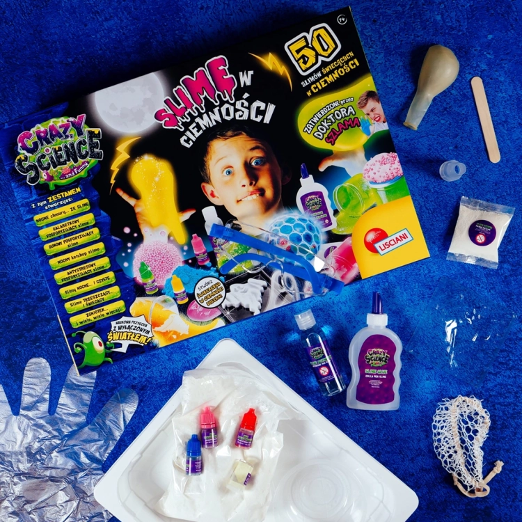 Pudełko zestawu Crazy Science Slime w ciemności oraz zawartość: klej, aktywator, barwniki, rękawiczka i akcesoria na niebieskim tle.