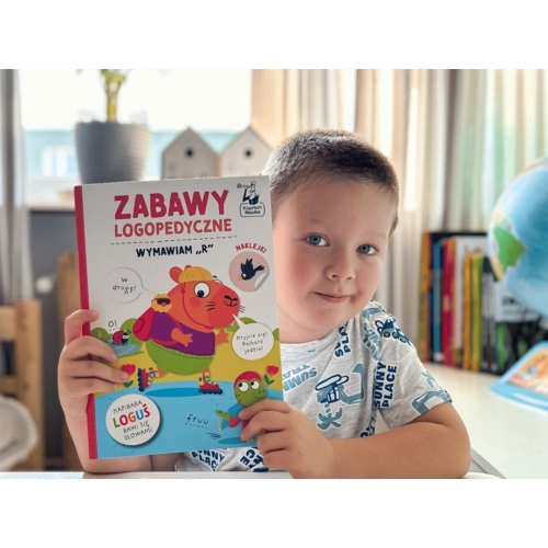 Chłopiec trzyma książkę Zabawy logopedyczne Kapibara Loguś Wymawiam R wydawnictwa Kapitan Nauka.