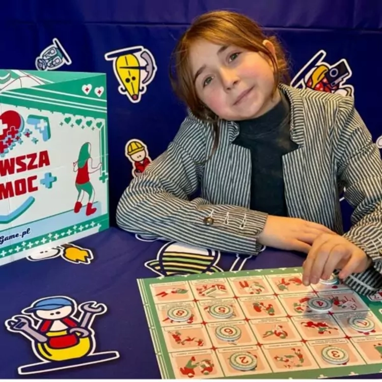 Dziewczynka prezentująca grę edukacyjną Pierwsza Pomoc BeGame 10+ przy stole z planszą i żetonami.
