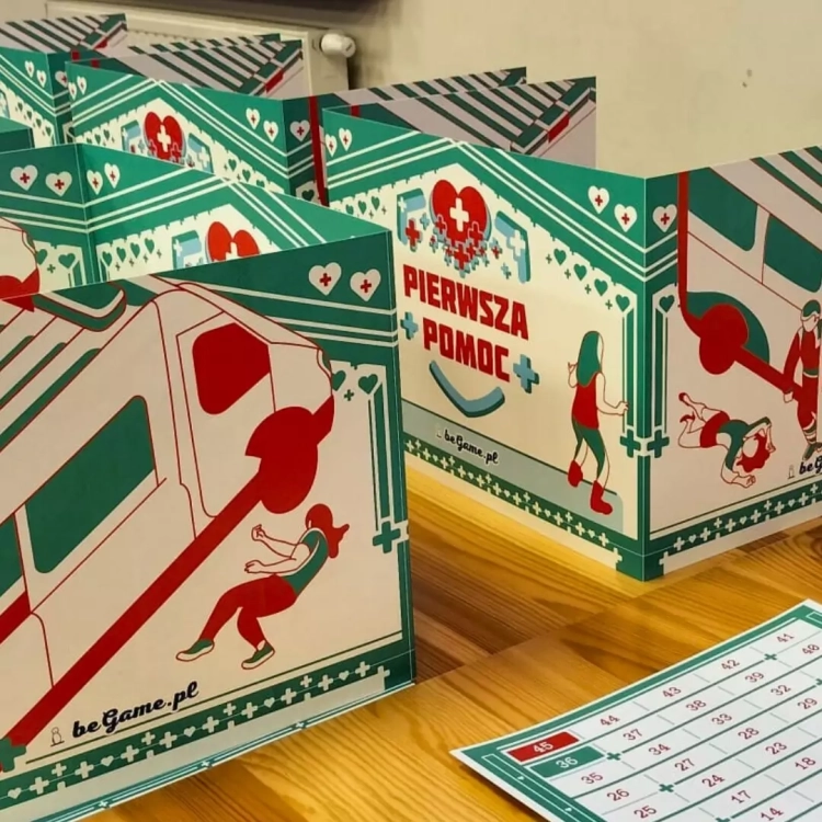 Parawany graczy z grafiką ambulansu i logo gry edukacyjnej Pierwsza Pomoc BeGame na drewnianym stole.