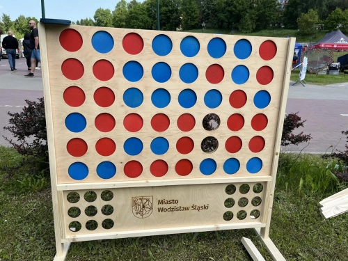 Drewniana gra "Kółko i krzyżyk na cztery" (Connect Four) w wersji XXL ustawiona w parku. Na planszy znajdują się niebieskie i czerwone krążki, a na dole widnieje napis "Miasto Wodzisław Śląski".
