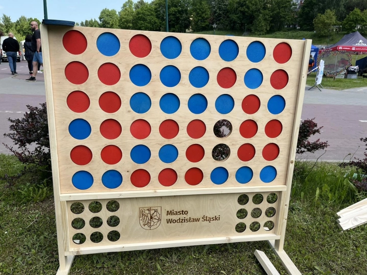 Drewniana gra "Kółko i krzyżyk na cztery" (Connect Four) w wersji XXL ustawiona w parku. Na planszy znajdują się niebieskie i czerwone krążki, a na dole widnieje napis "Miasto Wodzisław Śląski".