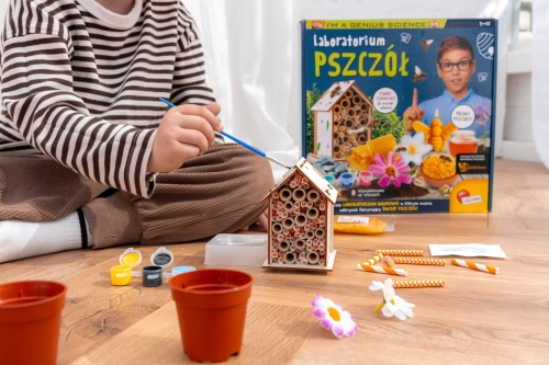 Dziecko siedzące na podłodze maluje pędzelkiem dach złożonego, drewnianego domku dla pszczół. Obok leżą farbki, doniczki i ozdobne kwiatki, a w tle stoi pudełko z zestawem.