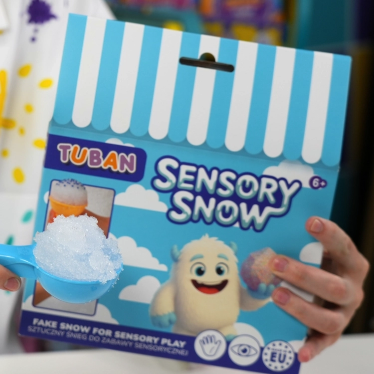 Niebieska łopatka pełna sztucznego śniegu na tle kolorowego opakowania zestawu Sensory Snow marki Tuban trzymanego w dłoni.